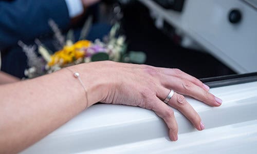 Detalle del anillo de recién casada en la mano de la novia.
La mano esta sobre el marco de la ventanilla de un coche clásico blanco.